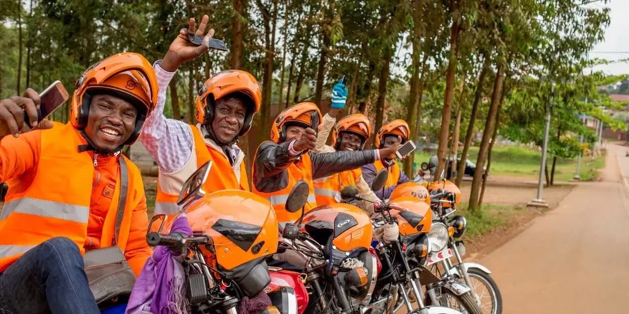 Boda riders in orange helmets and vests lined up on their bikes, smiling and holding up phones