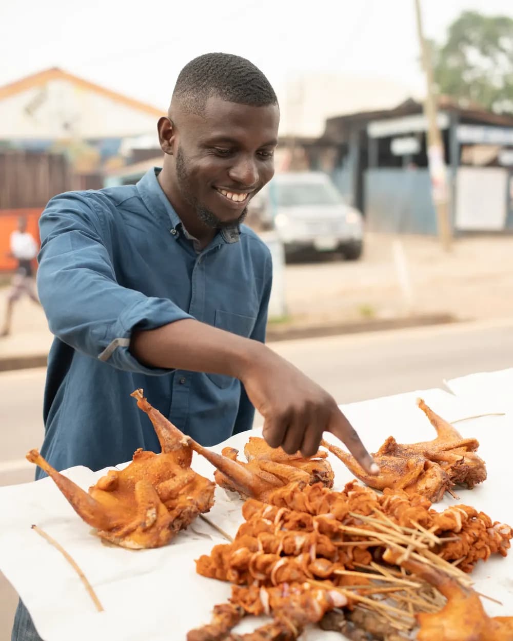 Street food vendor smiling over a roadside table of grilled chicken and skewers